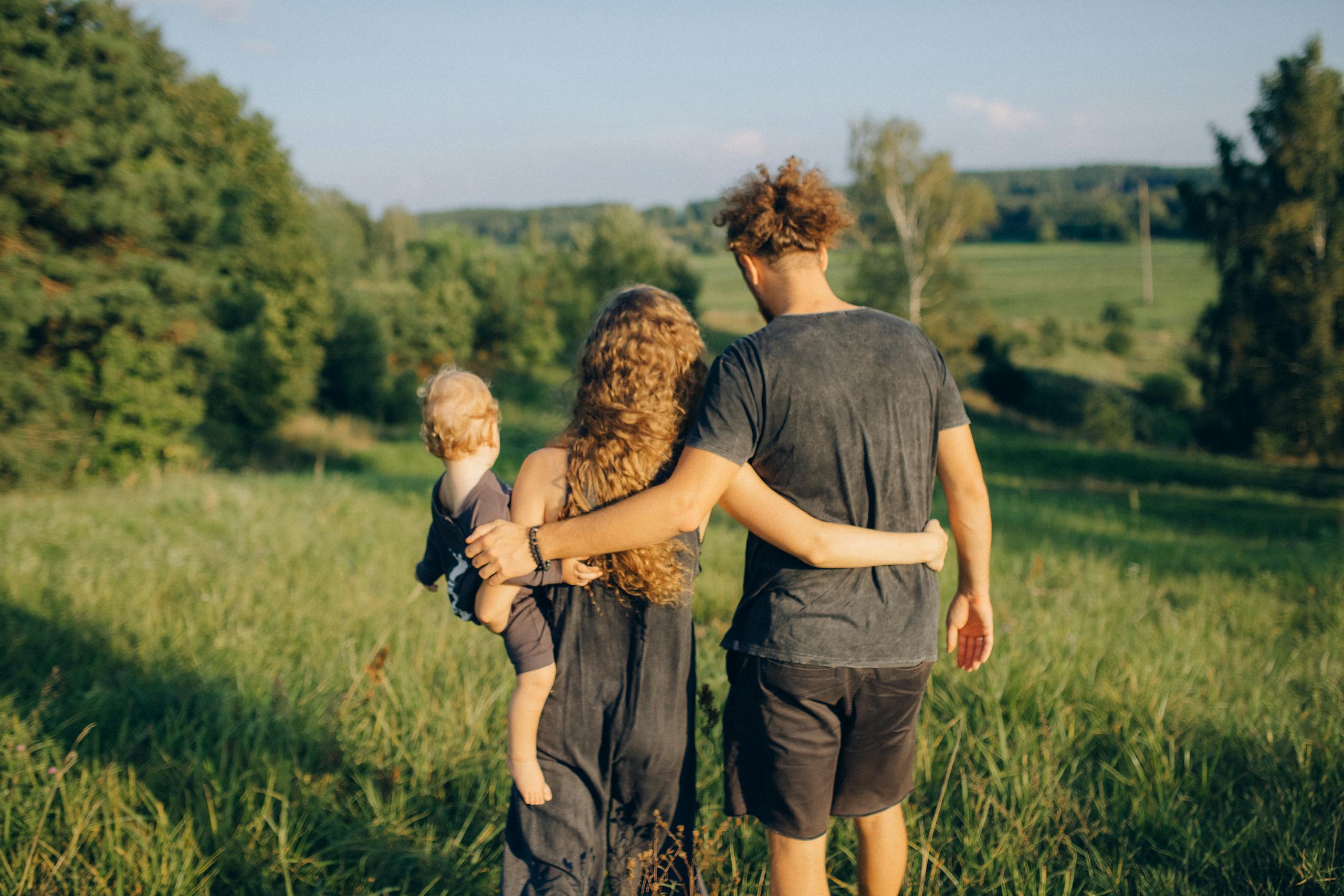 A couple with their child enjoy a serene walk through a sunlit meadow, surrounded by nature.