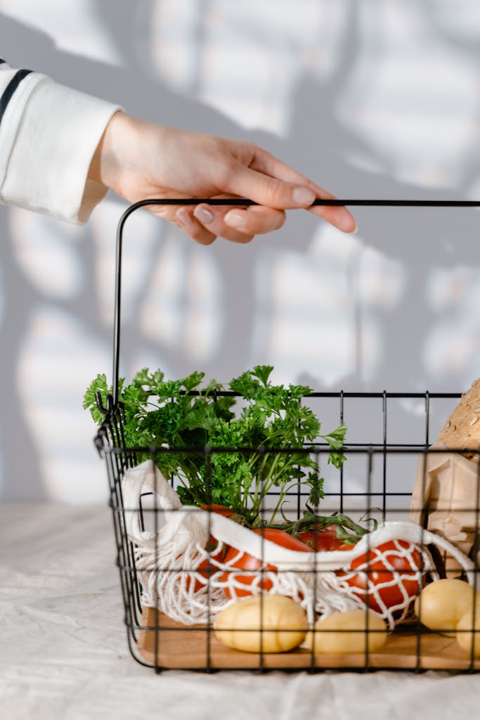 A hand holding a metal basket containing fresh organic vegetables like tomatoes and parsley.