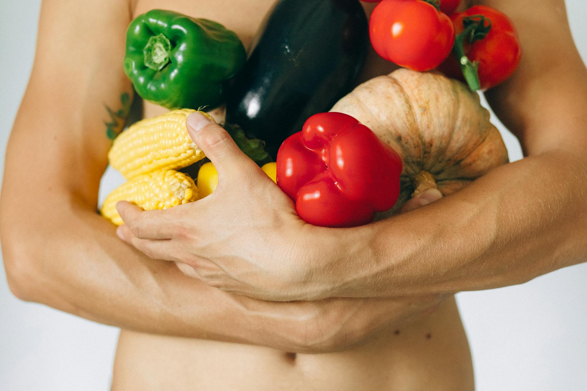 A man's torso holding a variety of fresh vegetables including peppers and corn.