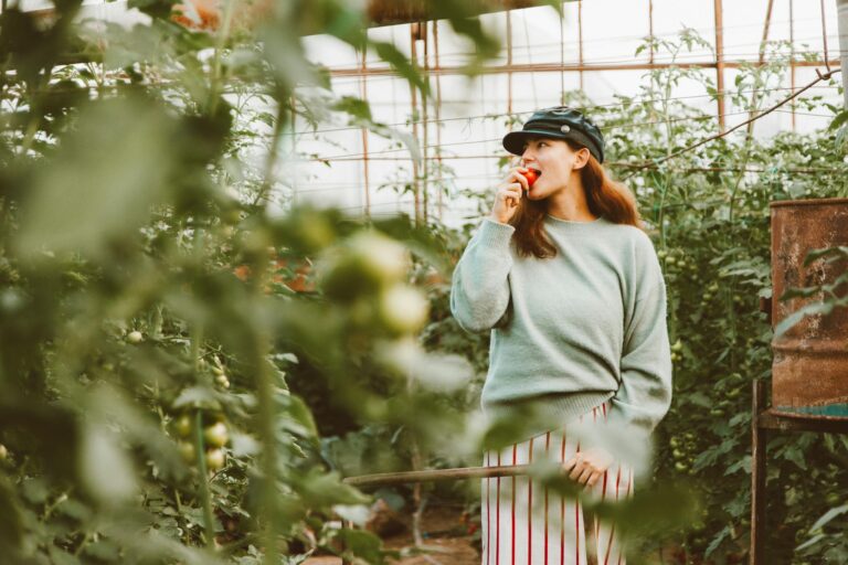 Adult woman enjoying a fresh tomato in a lush greenhouse setting.